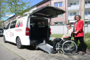 Ein Mann im Rollstuhl wird von einer Frau zu einem barrierefreien Fahrzeug mit geöffneter Heckrampe geschoben.